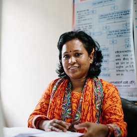 A woman in a bright orange sari sits at a desk and looks at the camera