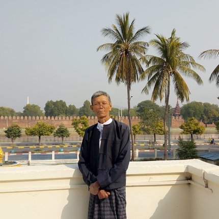 A man from Myanmar in dark clothes stands on a balcony overlooking a road with palm trees