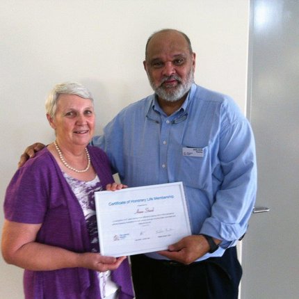 A woman with white hair in a purple cardigan receives a certificate from the now former TLM Australia Country Leader