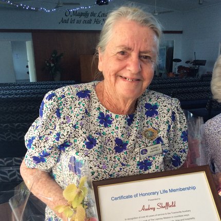 A woman with grey hair in a white and blue flowery dress holds up a commemorative certificate and a bouquet of flowers