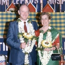 A woman and man in smart dress wear flower garlands at an event