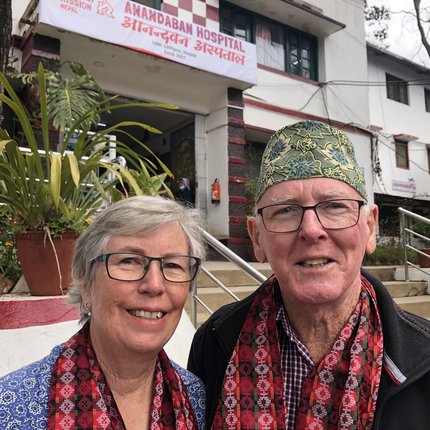 A white Australian couple wearing some traditional Nepali items (a hat and scarves) stand in front of Anandaban Hospital in Nepal
