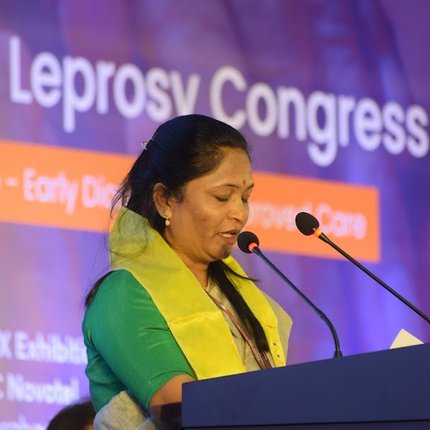 A women from India in bright yellow/green clothes speaks into a podium microphone