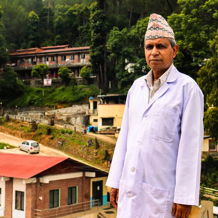 Dr Mahesh stands in front of Anandaban Hospital in a doctor's jacket and a traditional Nepali hat