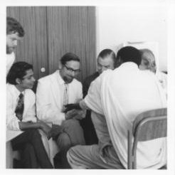 A black and white photo of a doctor with dark hair examining a patient as trainees watch