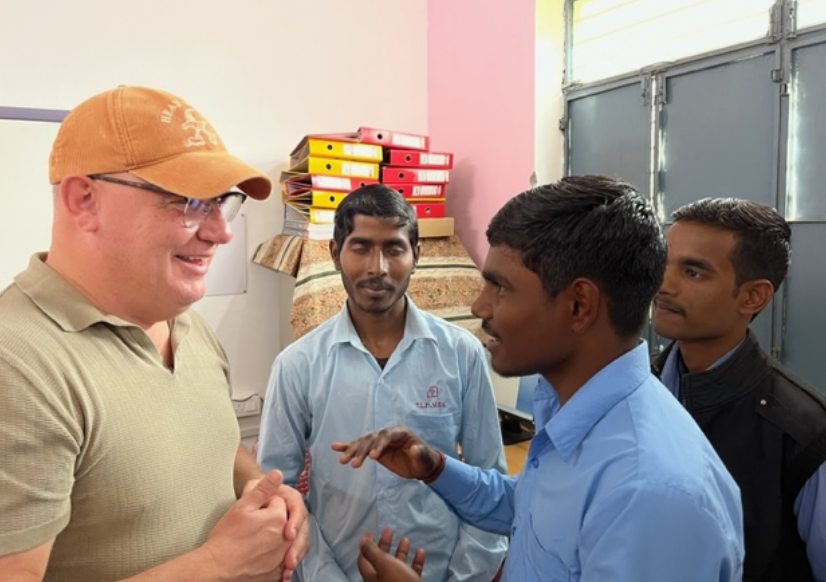 A Danish man in an orange baseball cap, glasses, and a green t-shirt speaks with three men from India who are wearing blue shirts