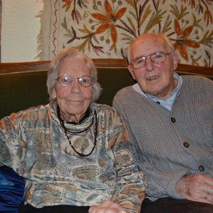 An older German couple in glasses sit together looking at the camera