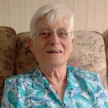 A woman in glasses with white hair smiles at the camera from a sofa