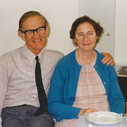 A man in a shirt and tie and black-rimmed glasses smiles while he puts his ram around his wife, who is wearing a pale coloured dress and blue cardigan