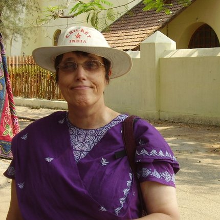 A white British woman in a cricket hat, tinted glasses, and a purple sari smiles to the camera