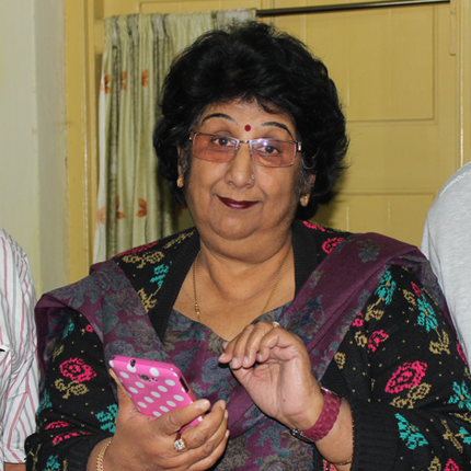 An Indian woman with short hair and glasses holds a phone and looks at the camera