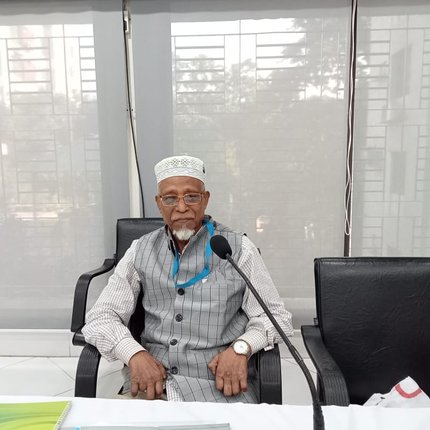 A man from Bangladesh in a white hat and grey suit sits behind a microphone at a desk