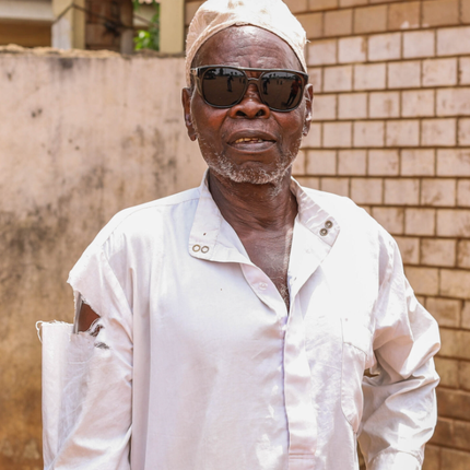 A Nigerian man in a white hat and clothes. He wears black sunglasses and has a small white beard