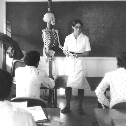 A black and white photo of a woman in front of a chalk board as she teaches a class of adult students