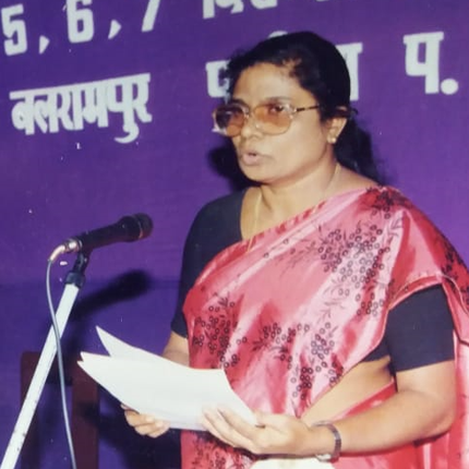 A woman in a pink saree and glasses speaks in front of a microphone