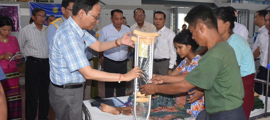 A representative of TLM Myanmar and another man hold a pair of crutches in front of a group of people, while a woman lies in a bed.
