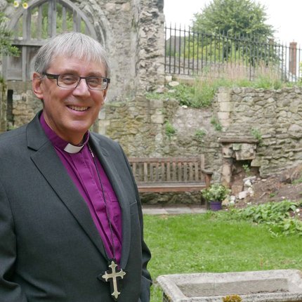 A white, middle-aged man in a purple Bishop's shirt smiles at the camera