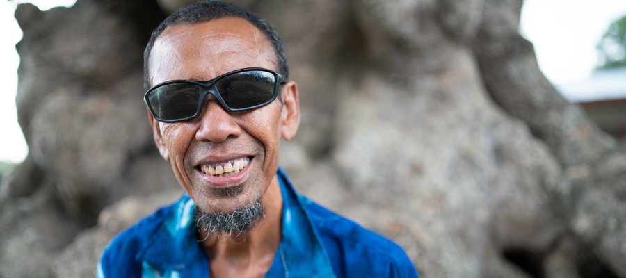 A man from Papua New Guinea wearing a blue shirt and sunglasses smiles at the camera