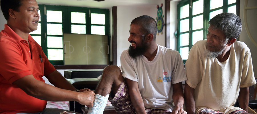 One man smiles at another man while he applies a dressing to his injured foot