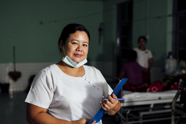 A nurse dressed in white blouse and red skirt lowers her face mask to pose for a picture at our partner hospital in Myanmar