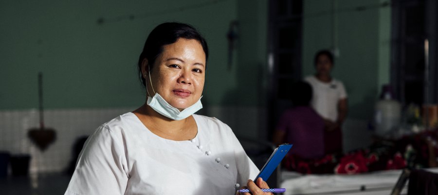 A nurse dressed in white blouse and red skirt lowers her face mask to pose for a picture at our partner hospital in Myanmar