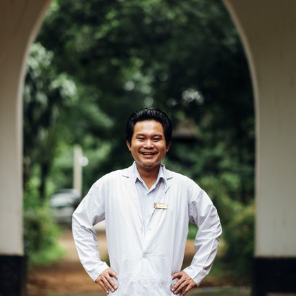 A man from Myanmar stands in a doctor's coat, smiling with hands on his hips