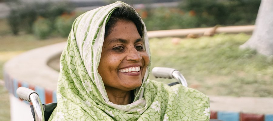 A woman sat in a wheelchair smiling at the camera. Her head is covered with a shawl.