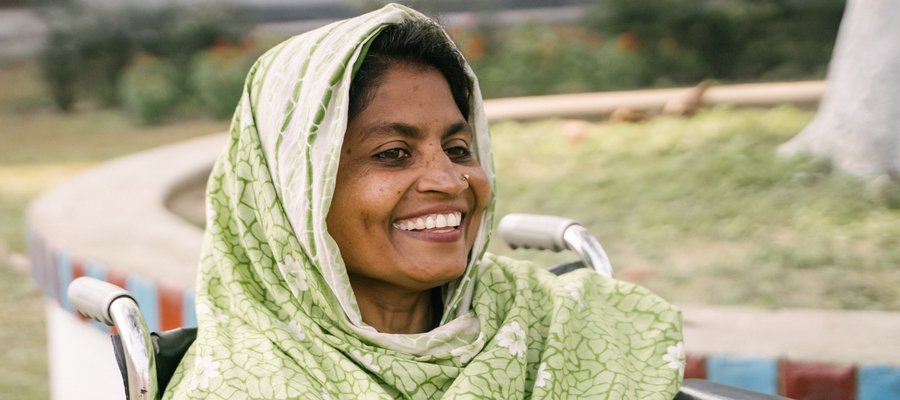 A woman sat in a wheelchair smiling at the camera. Her head is covered with a shawl.