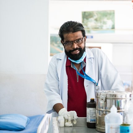 A Nepali man in a doctor's outfit on a hospital ward smiles at the camera