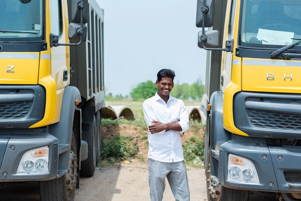 A young Indian man in a white shirt stands between two yellow lorries smiling