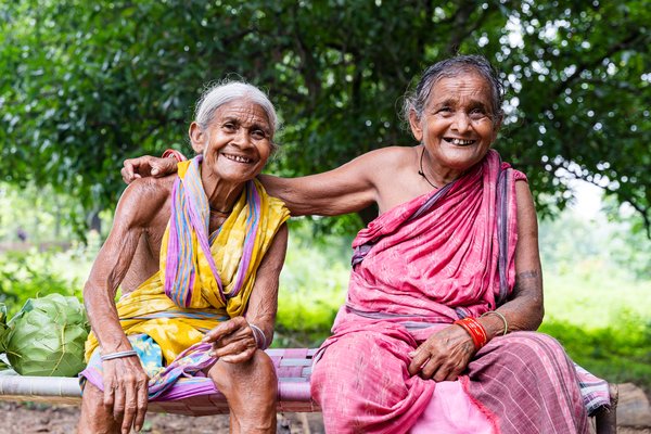 Two older ladies from India sit on a bench, one has her arm around the shoulders of the other