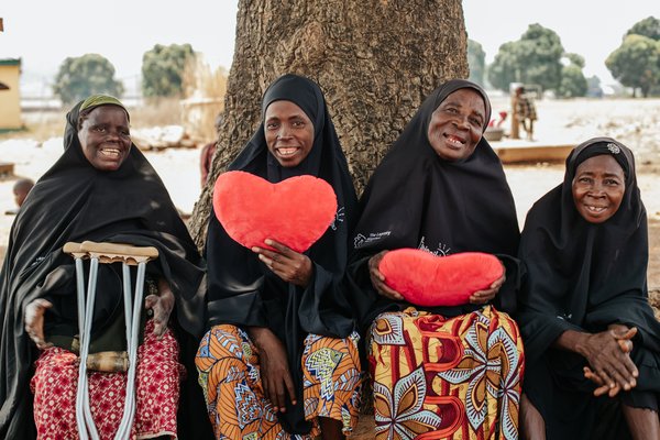 Four Nigerian women wearing black sit on a bench, two of whom hold up red hearts