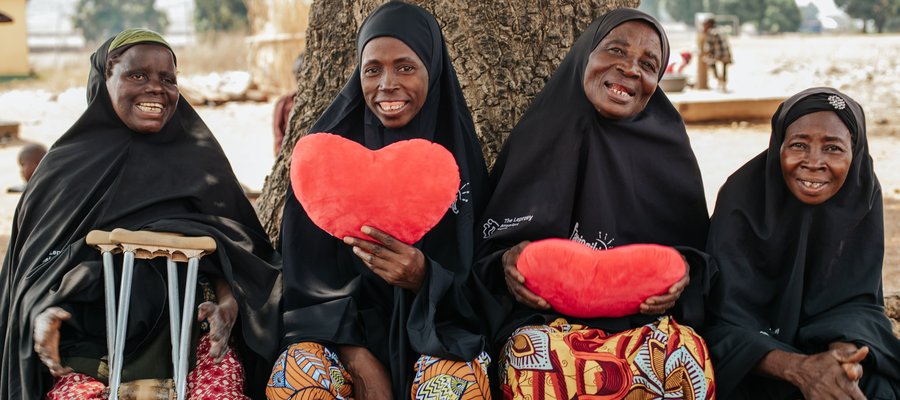 Four Nigerian women wearing black sit on a bench, two of whom hold up red hearts