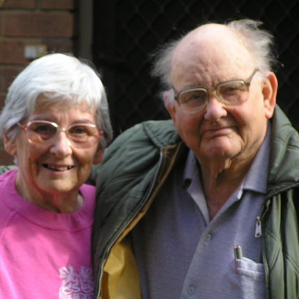 An older couple look at the camera with their arms around each other outside a brick building