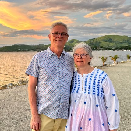 An older couple pose for a photo on the beach with rolling hills and a sunset behind them