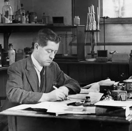 A black and white photo of a man in a suit with dark hair writing at a desk